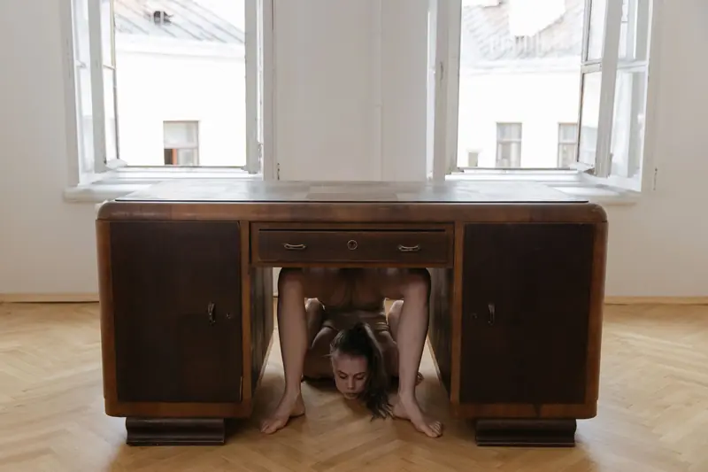 A young woman contortionist bends under a wooden desk in a minimalist interior setting