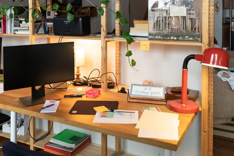 A cozy indoor workspace featuring a wooden desk, monitor, and red desk lamp, with plants and office supplies