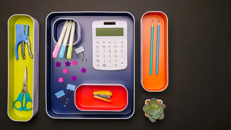 Flat lay of organized desk accessories in colorful trays including calculator, pens, and succulent plant