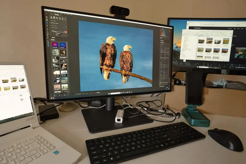A desk setup showing photo editing software on multiple monitors, a keyboard, and peripherals